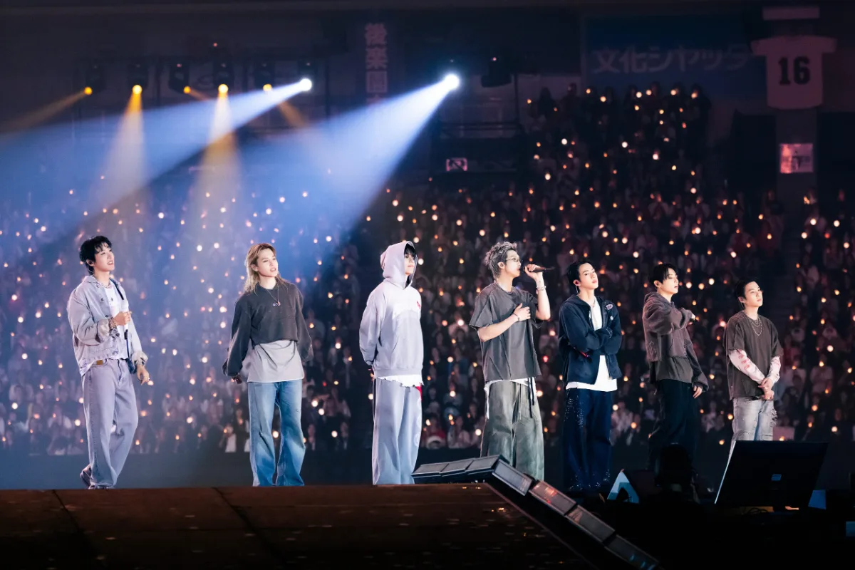 BTS no palco com a plateia iluminada ao fundo, em foto oficial de divulgação da turnê ARIRANG